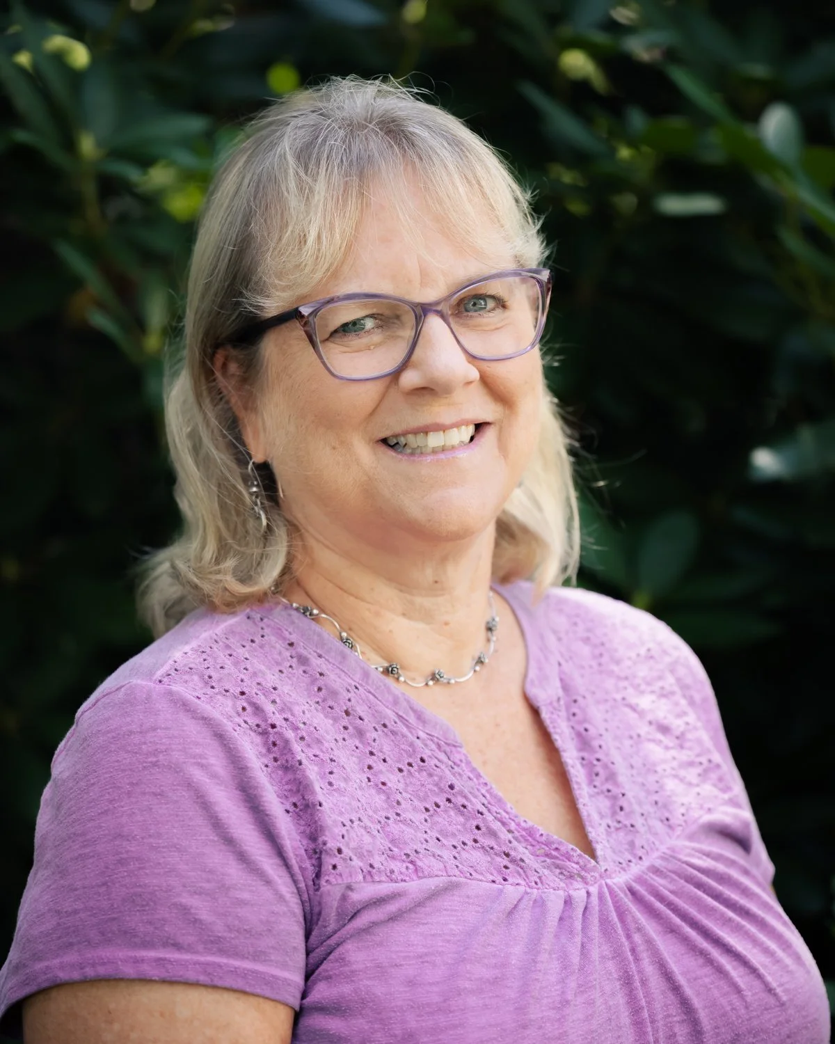 A smiling middle-aged woman with blonde hair, glasses, wearing a purple top and a necklace, standing outdoors with green foliage in the background.