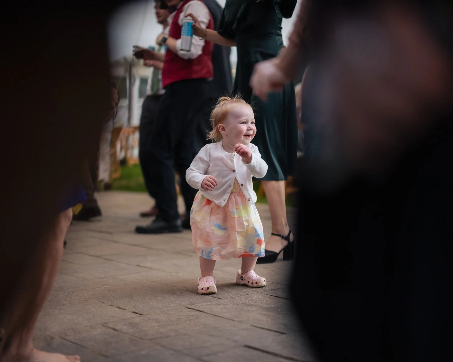 A young girl with blonde hair, wearing a white jacket, pastel dress, and pink Crocs, is smiling and walking on a wooden floor among adults at an indoor or outdoor social gathering.