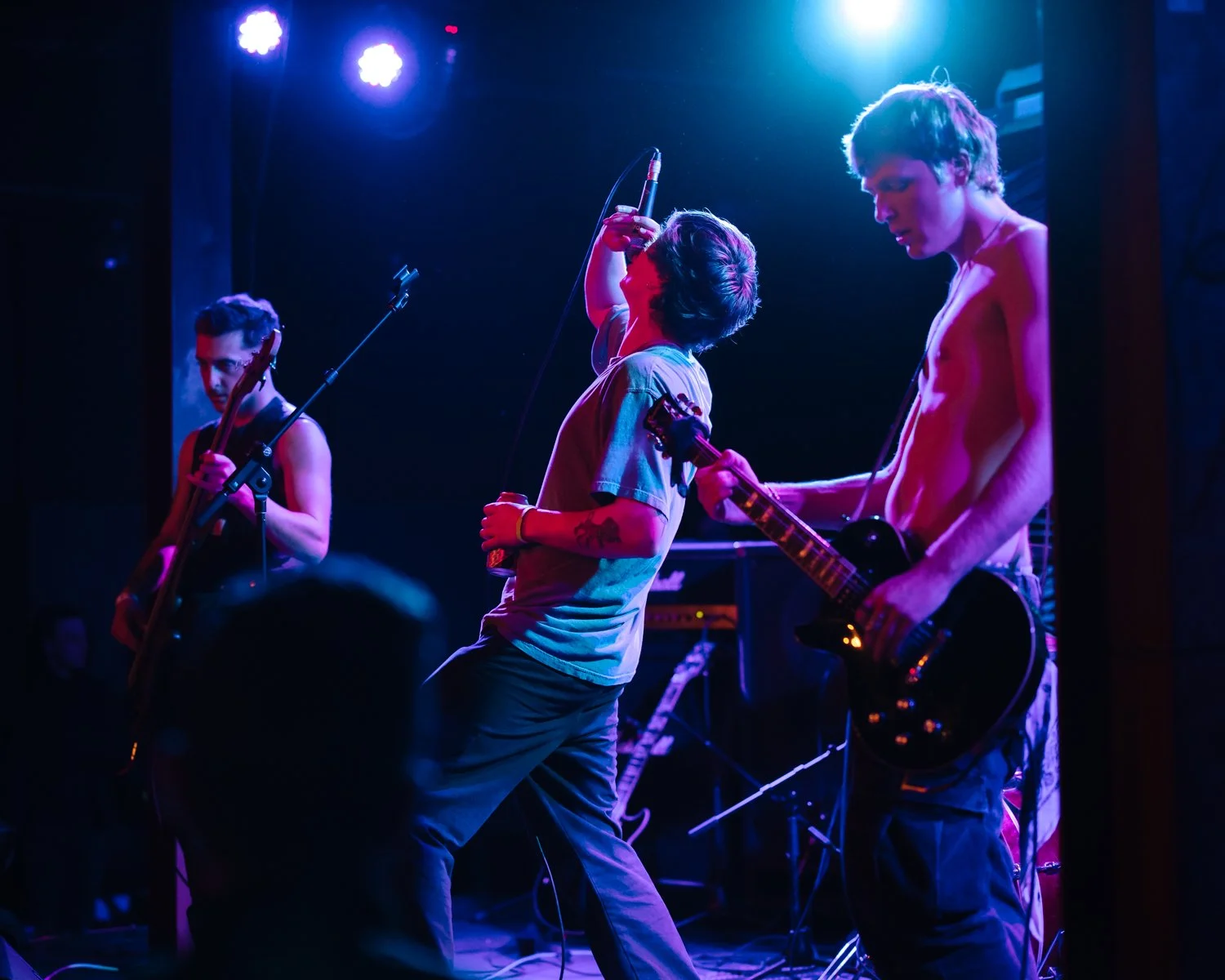 Three young men performing live music on stage, with two playing electric guitars and one singing into a microphone, illuminated by stage lights.
