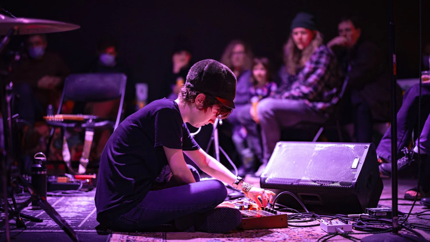 A young musician sitting on the stage floor, playing electronic music equipment, with an audience watching. Stage lighting creates a purple hue.