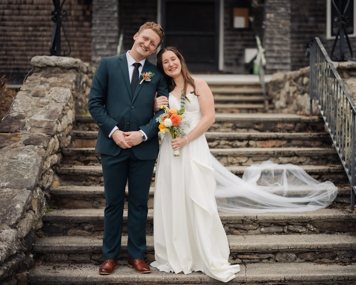 A smiling bride and groom standing on stone steps outside a building, with the bride holding a bouquet of orange, white, and yellow flowers. The groom wears a teal suit, and the bride wears a white wedding dress with a flowing veil on the ground.