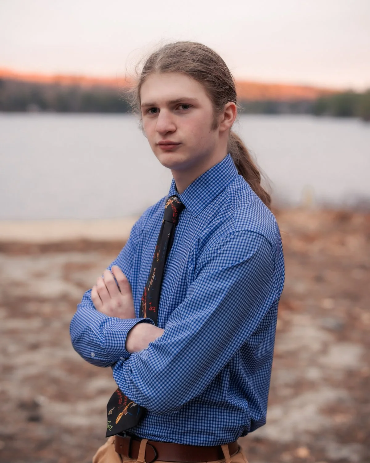 A young man with long hair tied back, wearing a blue checkered shirt and a tie, stands outdoors with arms crossed against a backdrop of a lake and a blurred landscape at sunset.