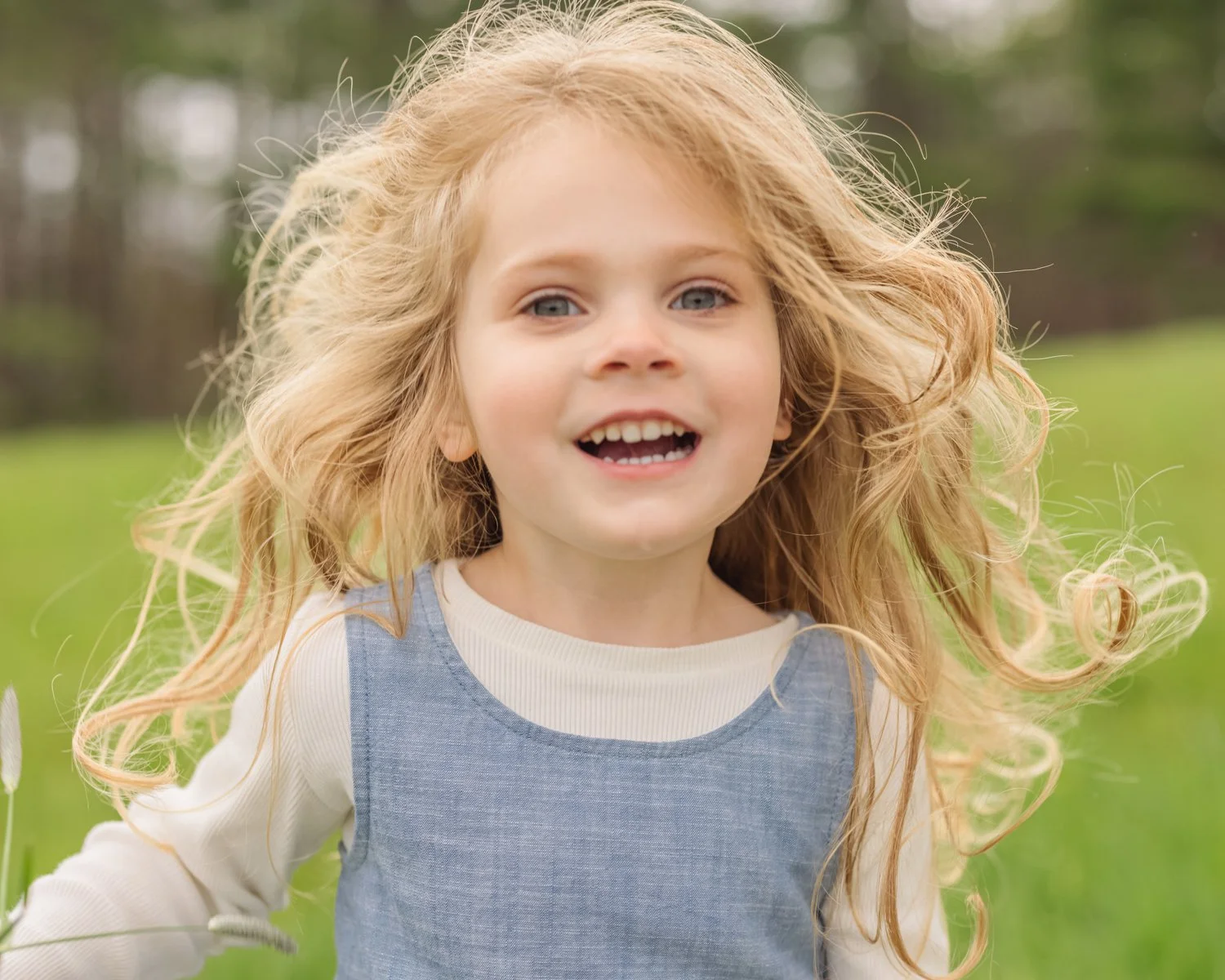 A young girl with long, wavy red hair and blue eyes smiling outdoors in a grassy field, wearing a light-colored long-sleeve shirt and a gray dress.