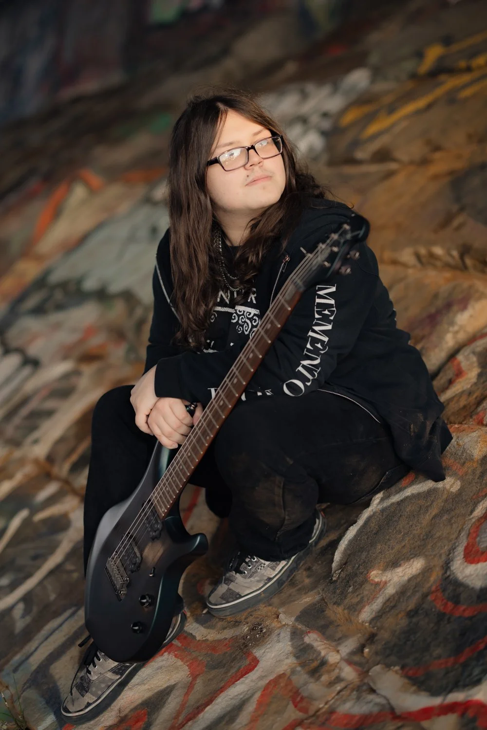 A young person with long brown hair, glasses, and a black jacket, sitting on a graffiti-covered wall while holding a black electric guitar.