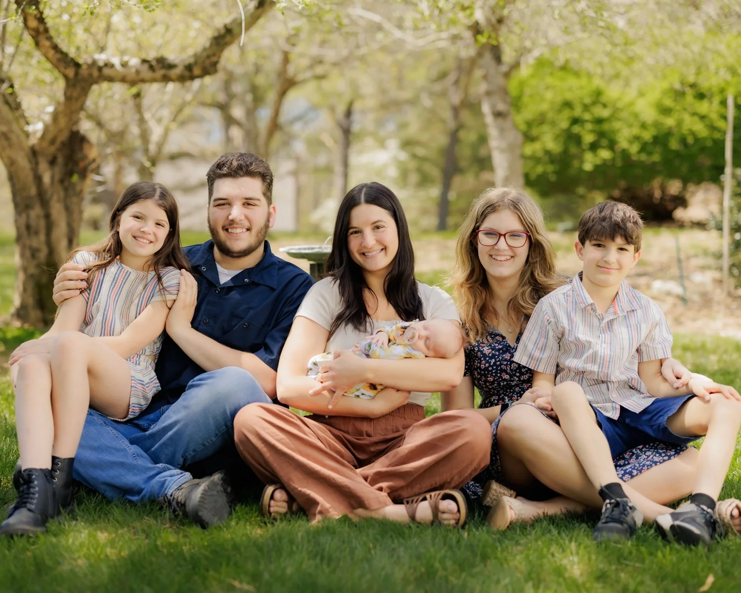 A family of five sitting on grass in a park with trees in the background, smiling at the camera. A woman in the center holds a baby, and two children sit on either side of her.