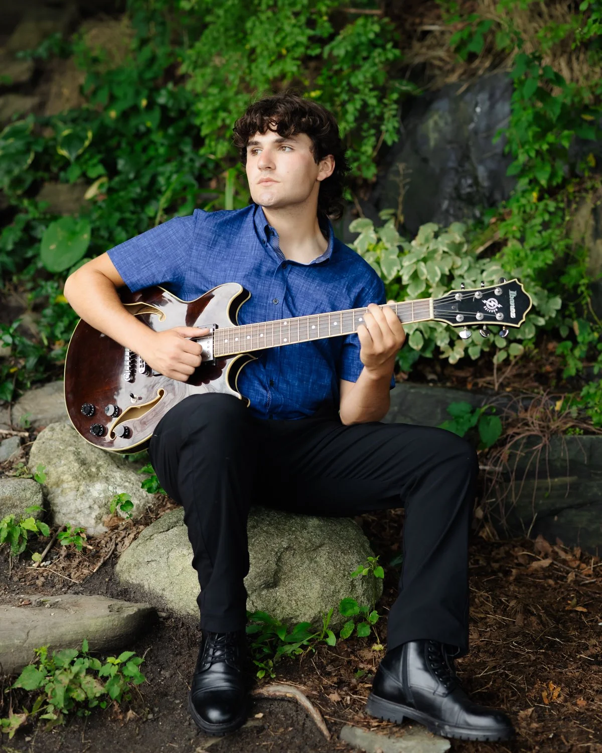 A young man with curly hair playing an electric guitar outdoors, seated on a large rock with greenery in the background.