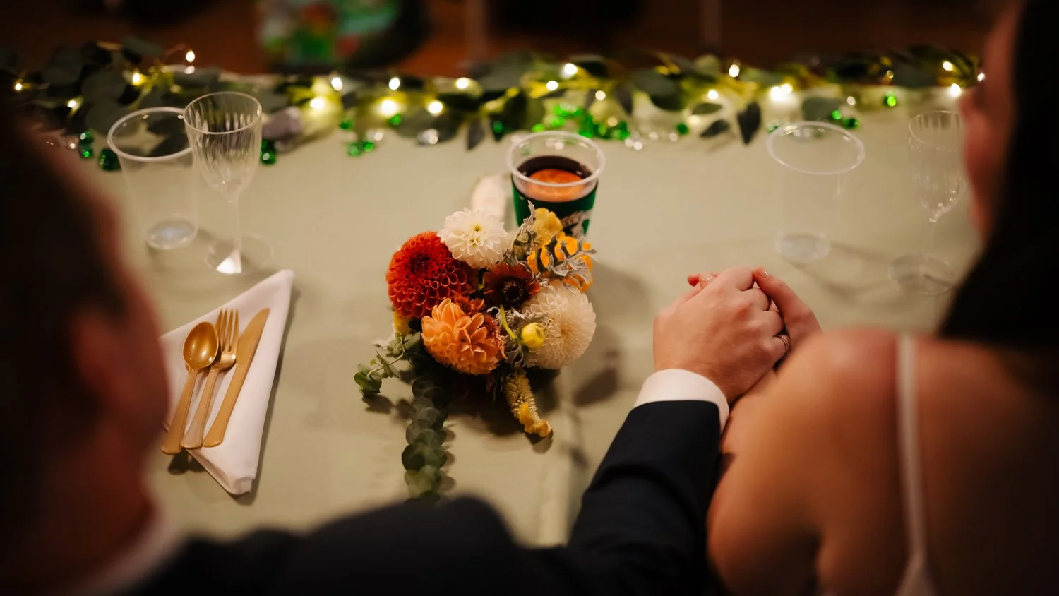 A couple holding hands at a decorated table with a flower arrangement and a drink, possibly at a wedding or celebration.