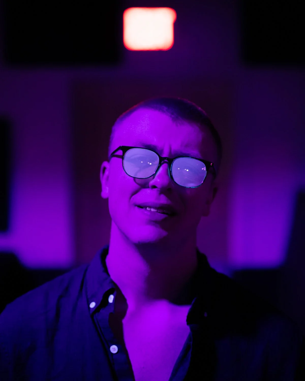A person wearing glasses with a colorful purple and pink lighting, in a dark room with an illuminated sign in the background.
