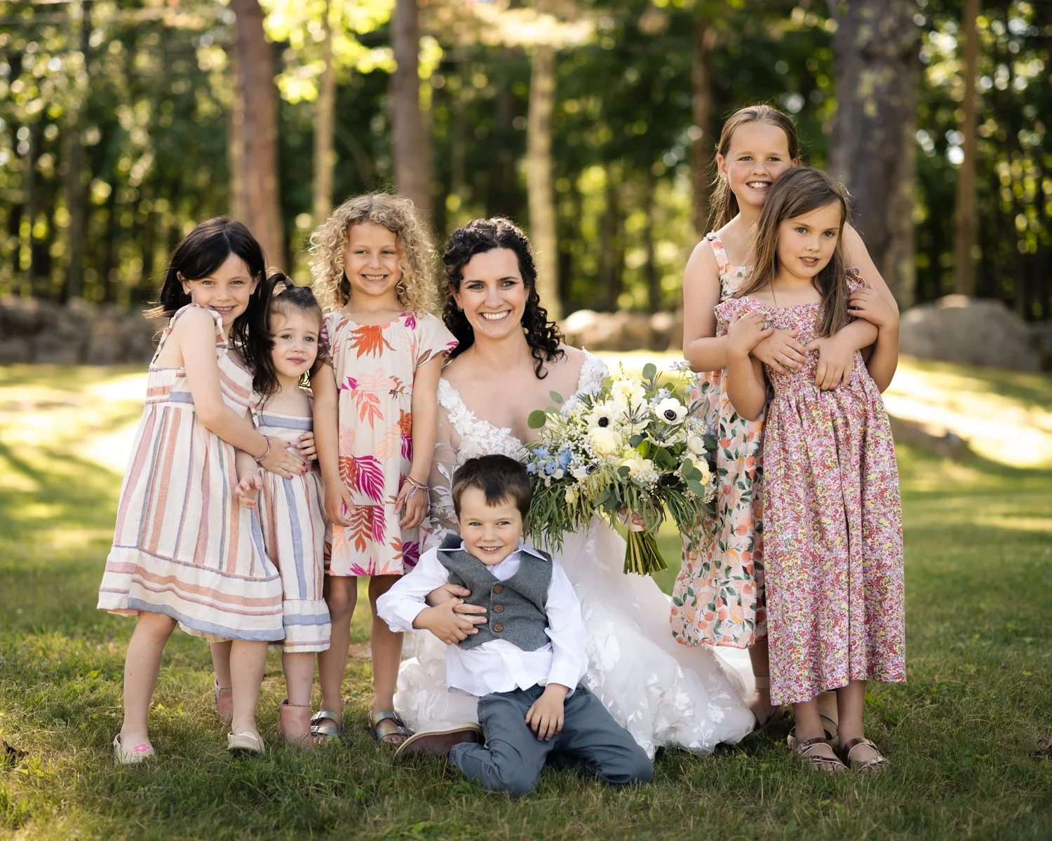A woman in a white wedding dress holding a bouquet of white and green flowers, surrounded by children in colorful dresses outside in a wooded park.