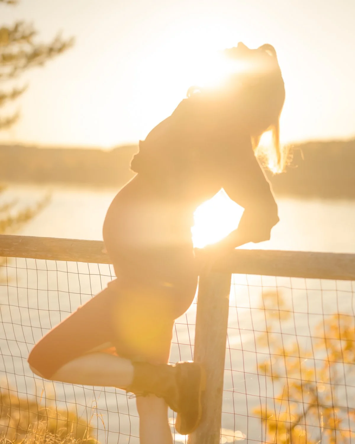 Silhouette of a girl climbing a wooden fence near a body of water during sunset.