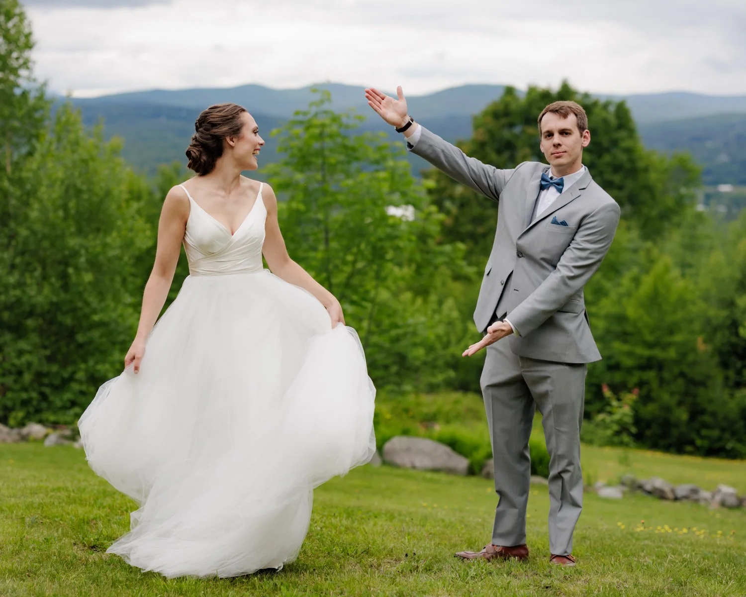 Bride in a white wedding dress smiling as she dances with a groom in a gray suit and blue bow tie outdoors with green trees and mountains in the background.