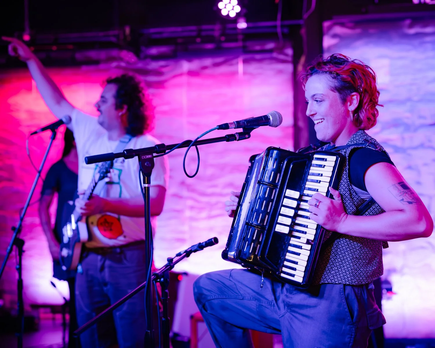 Musicians performing on stage with colorful lighting; woman playing an accordion and singing, man holding a guitar and singing in the background.