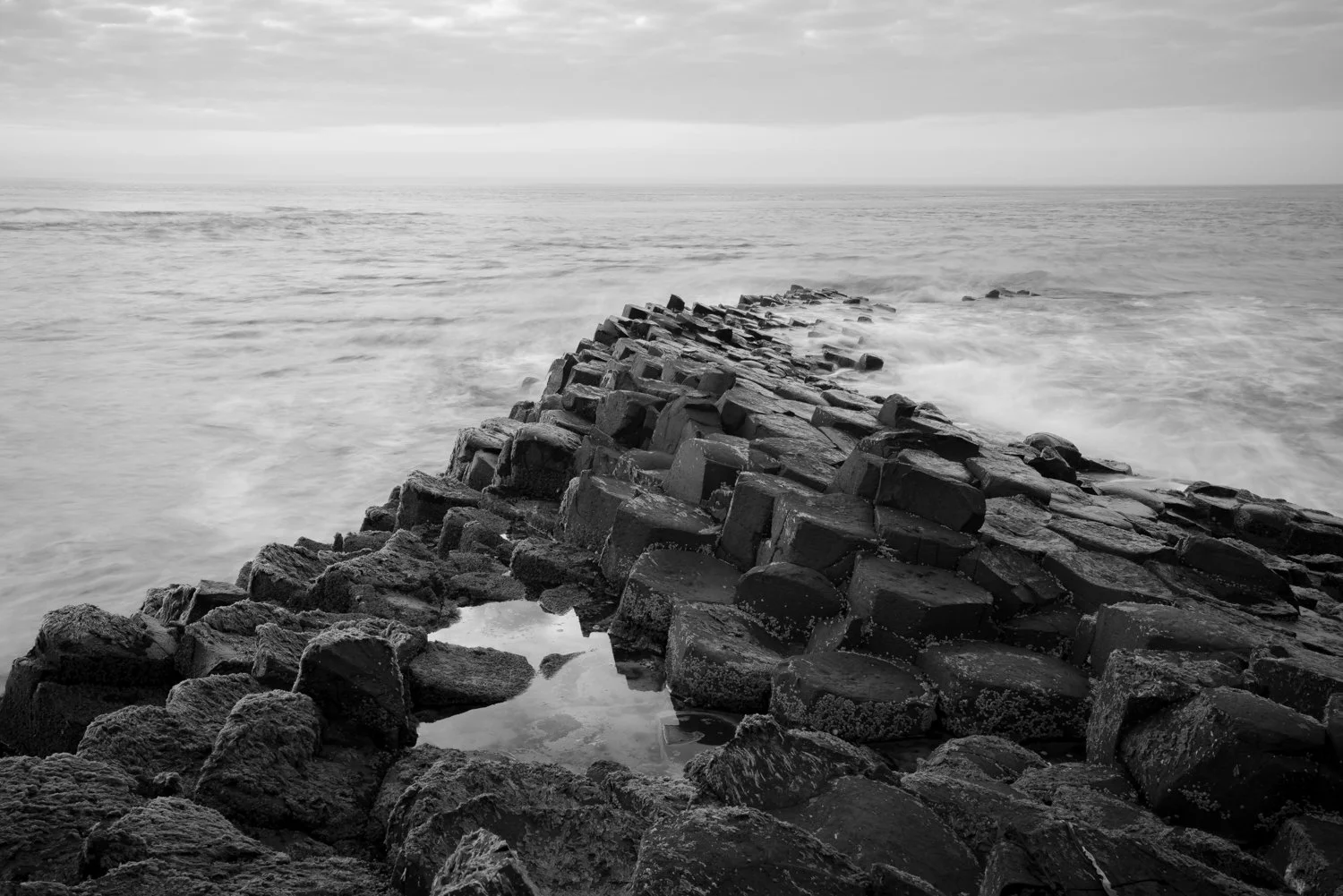 Black and white photo of a rocky pier extending into the ocean with waves crashing against it and a cloudy sky above.