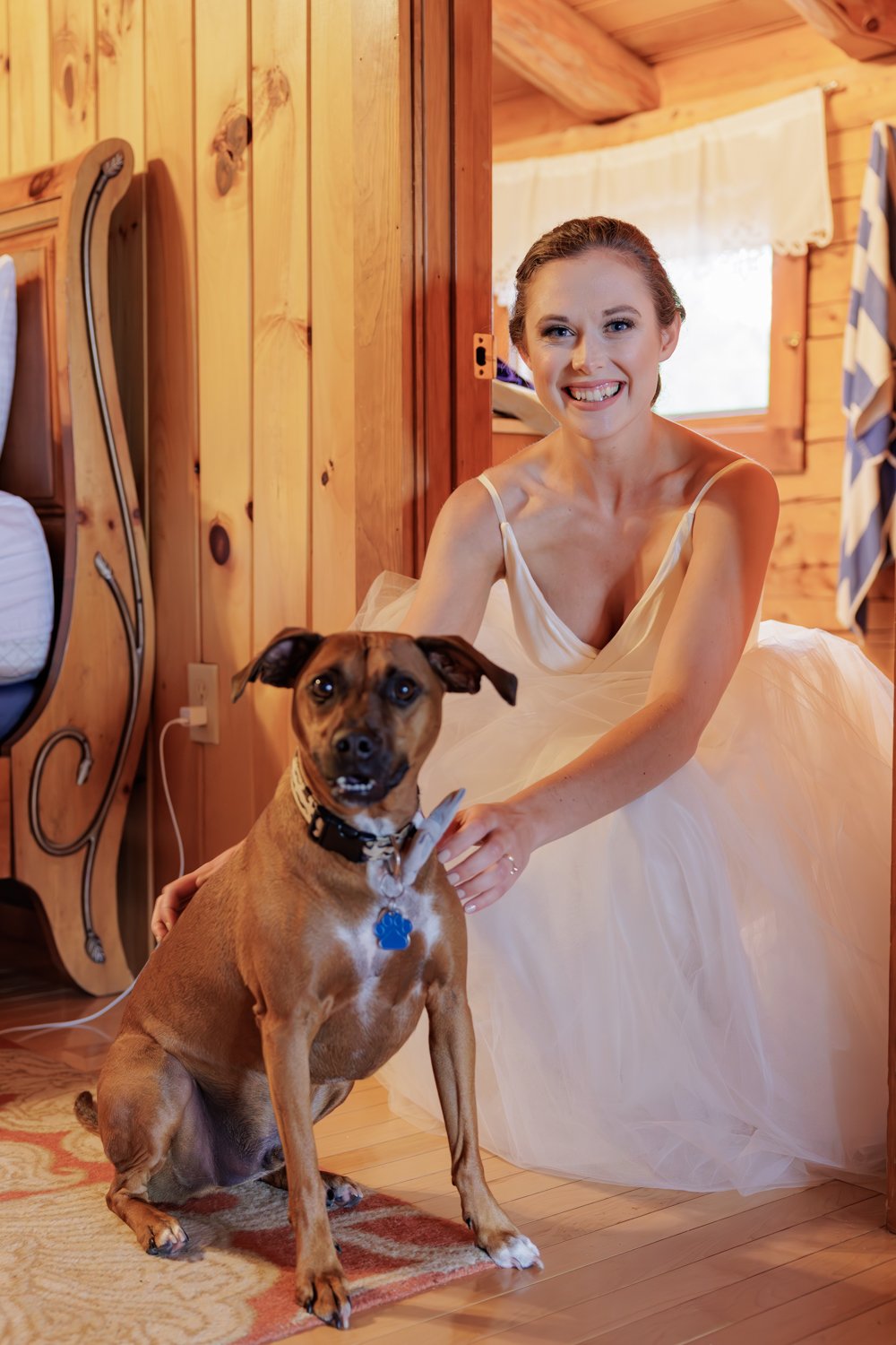 A woman in a white dress sitting on a wooden floor in a cabin, smiling and touching a brown dog with a black collar and a blue tag, in front of her.