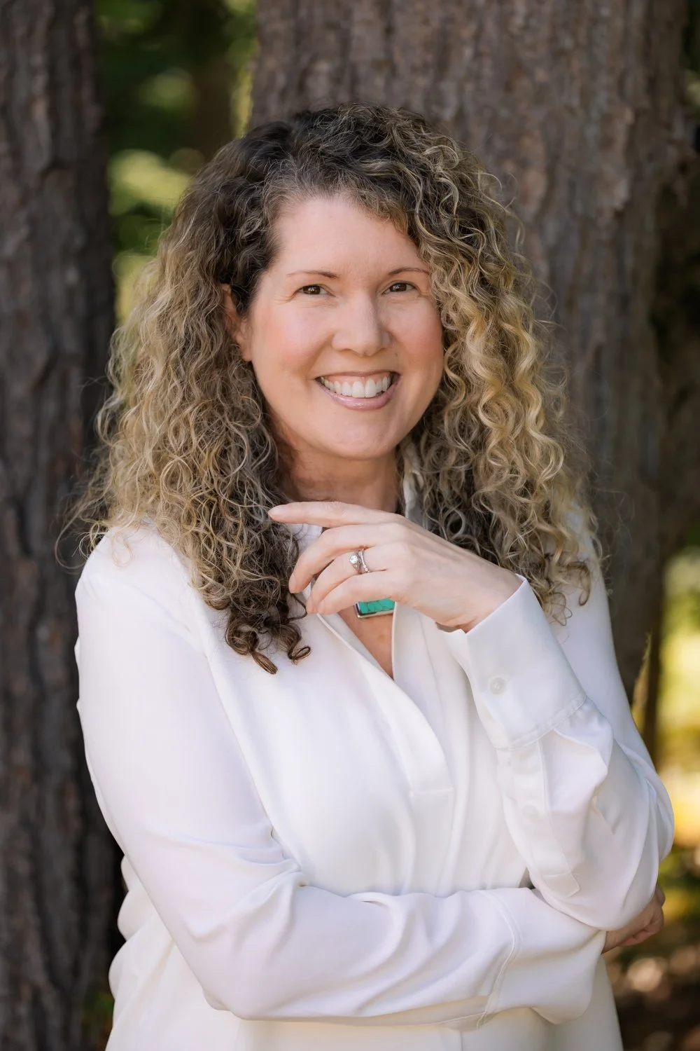 A woman with curly blonde hair smiling outdoors in front of a tree, wearing a white shirt and turquoise jewelry.