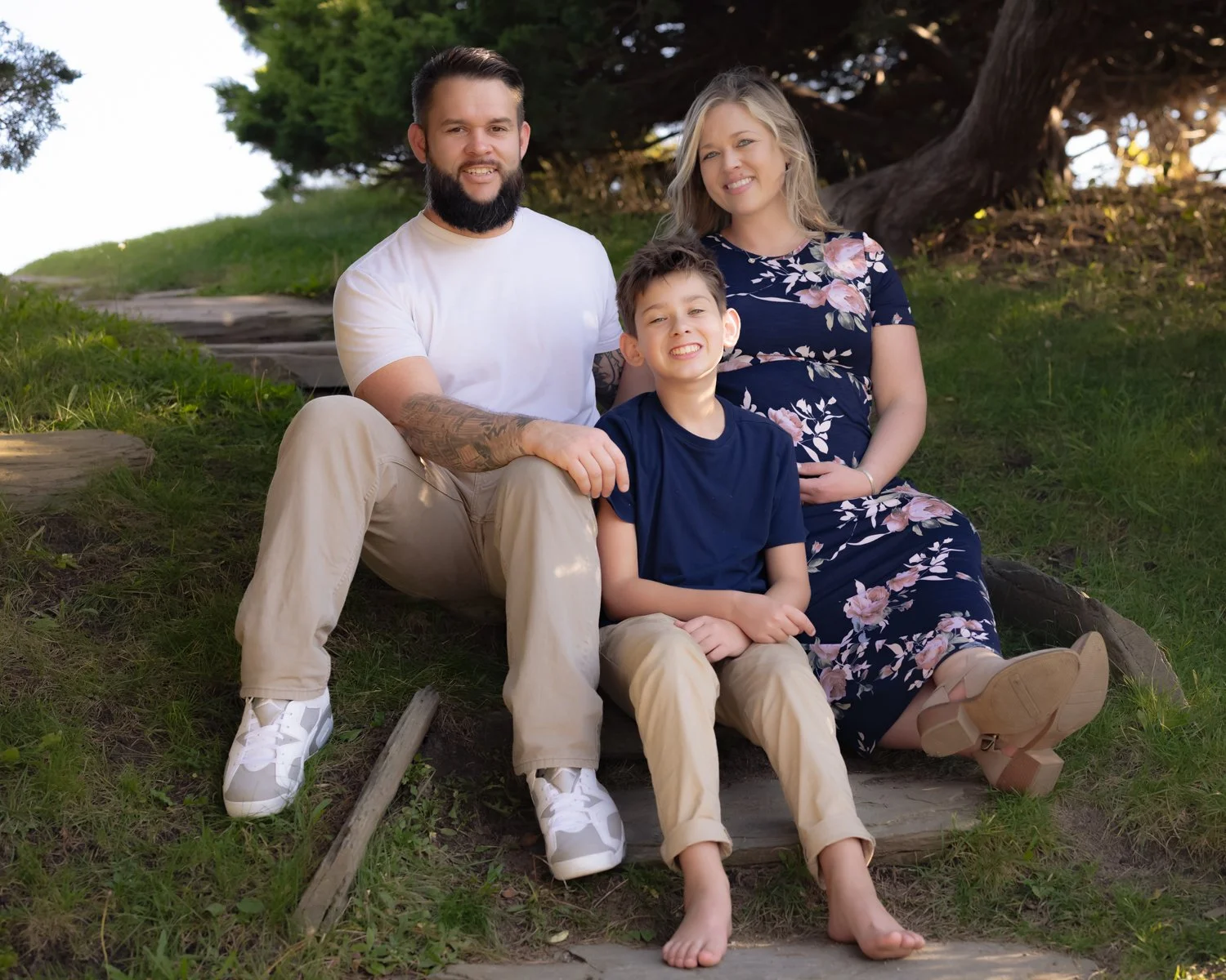 A family of four sitting outdoors on a grassy area near a tree, smiling at the camera. The father has a beard and is wearing a white t-shirt and beige pants. The mother has blonde hair and is wearing a floral dress. The son is in front, wearing a nav
