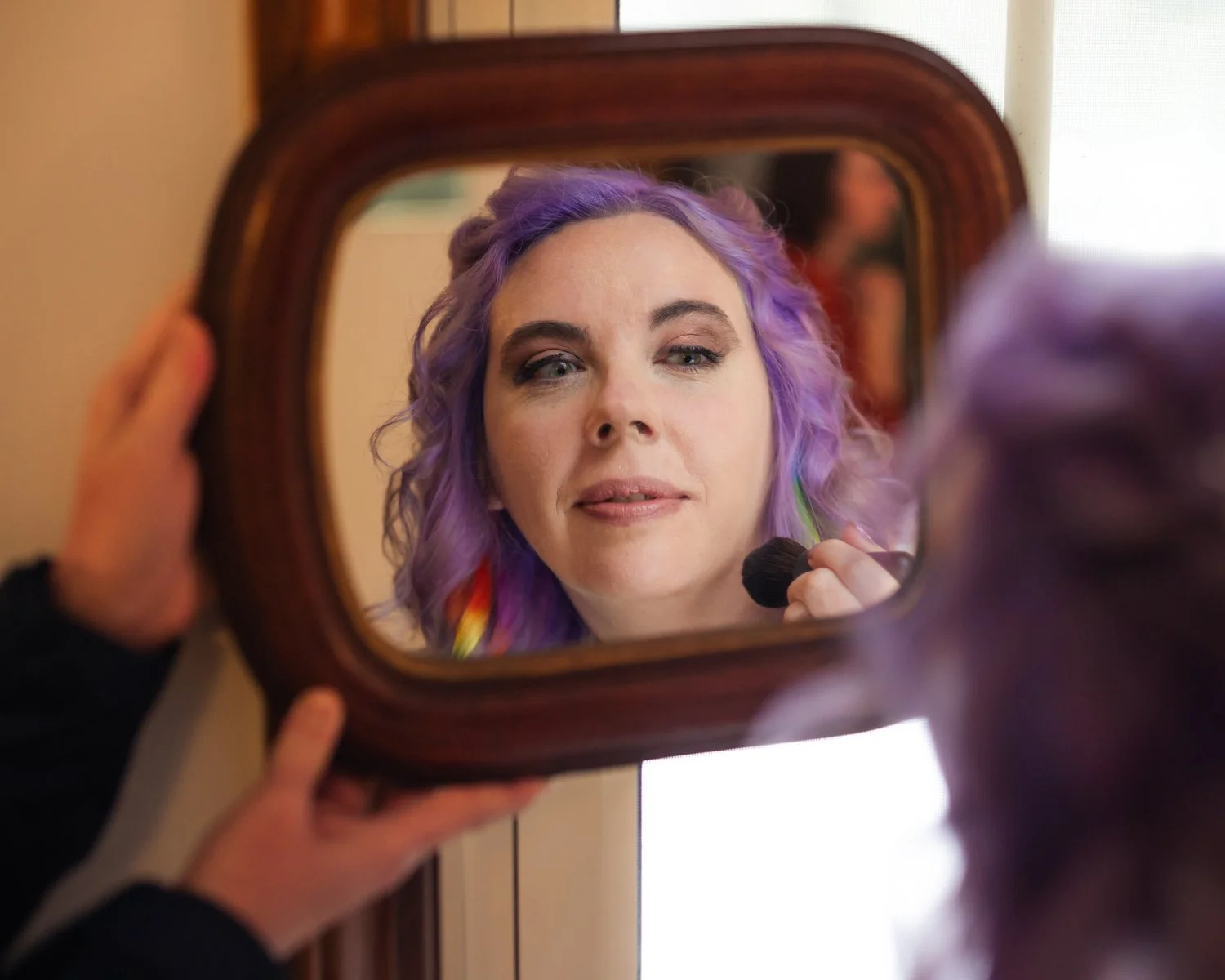 Woman with purple hair applying makeup in front of a small mirror.