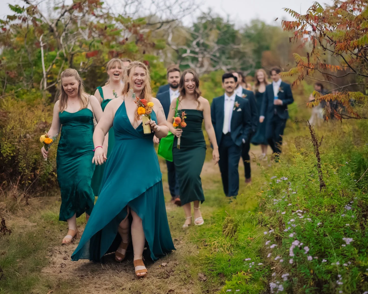 A group of people dressed in formal attire walking outdoors on a trail surrounded by greenery and trees with autumn leaves, smiling and laughing.