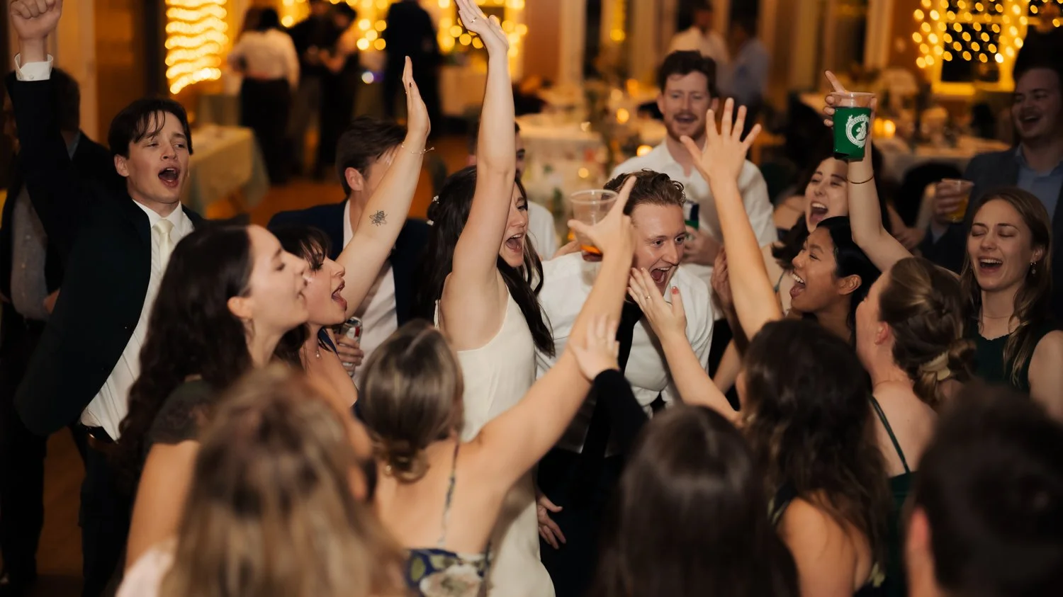 A group of people dancing and celebrating at a party or wedding reception, smiling, raising their hands, and holding drinks in a warmly lit venue.