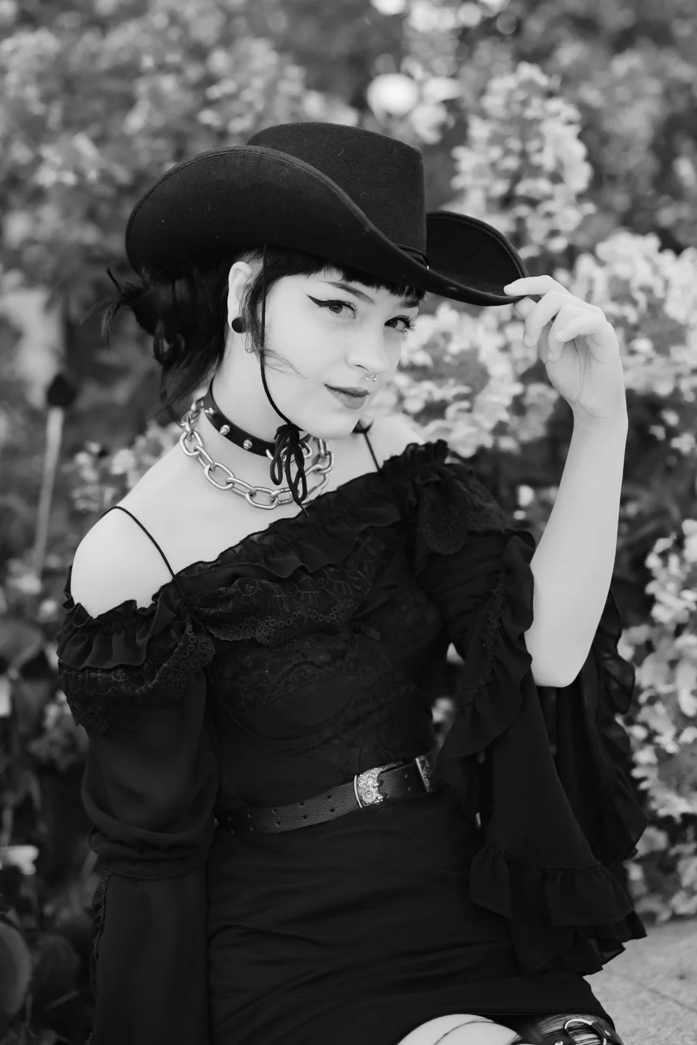 A woman with short dark hair and piercings, wearing a wide-brimmed hat, a chunky chain necklace, and a lace dress, sitting outdoors in front of flowers, holding the brim of her hat with one hand, smiling slightly.