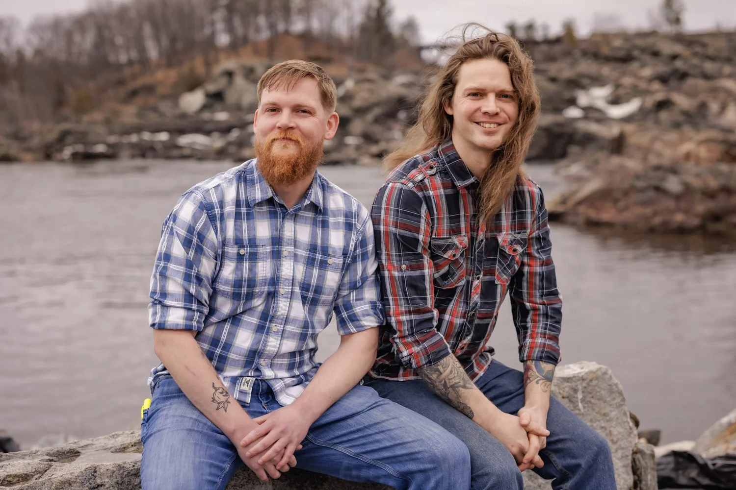 Two men sitting on rocks by a river, wearing plaid shirts, smiling, with trees and rocks in the background.
