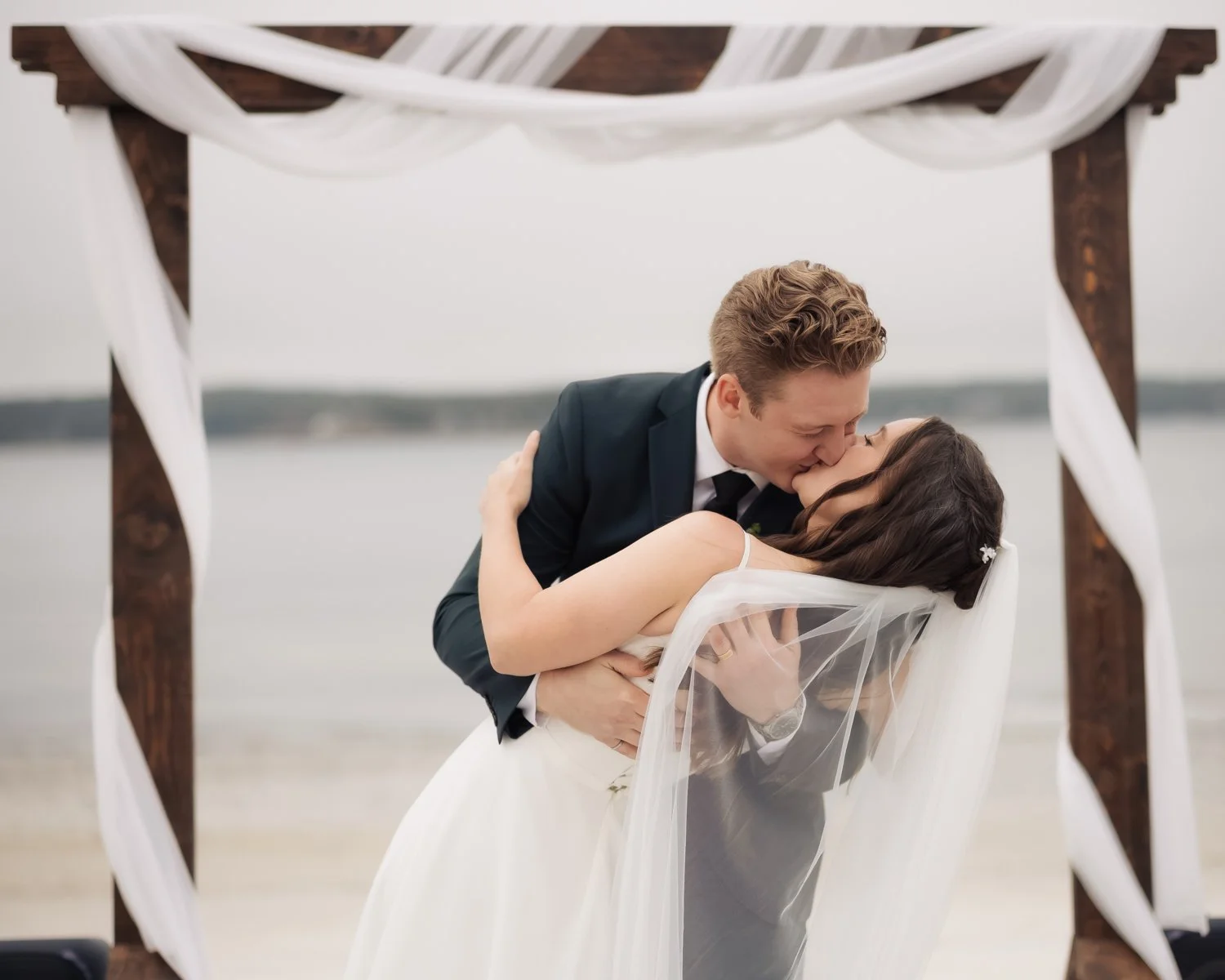 Man and woman kissing during wedding ceremony on the beach, framed by wooden arch with white drapes.