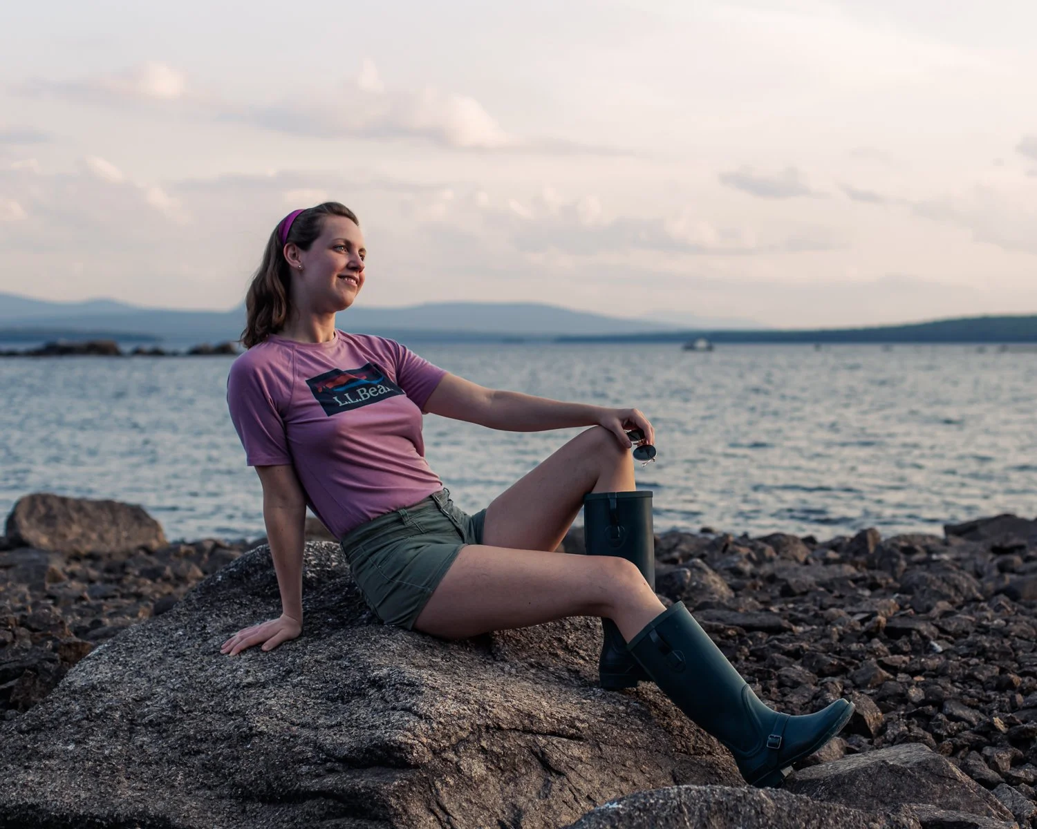 A woman in a pink L.L. Bean t-shirt and green shorts sits on a large rock by a body of water, holding a black thermos and smiling while looking into the distance. The background features a cloudy sky and distant mountains.