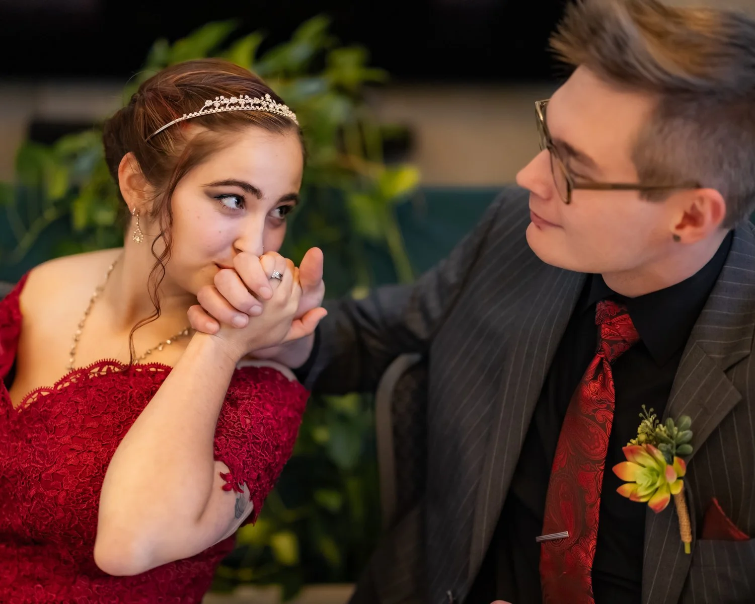 A young woman in a red lace dress is holding hands and kissing them with a young man in a dark striped suit, glasses, and a red tie, who is smiling and looking at her. The woman is wearing a tiara and jewelry, and the man has a boutonniere with a flo