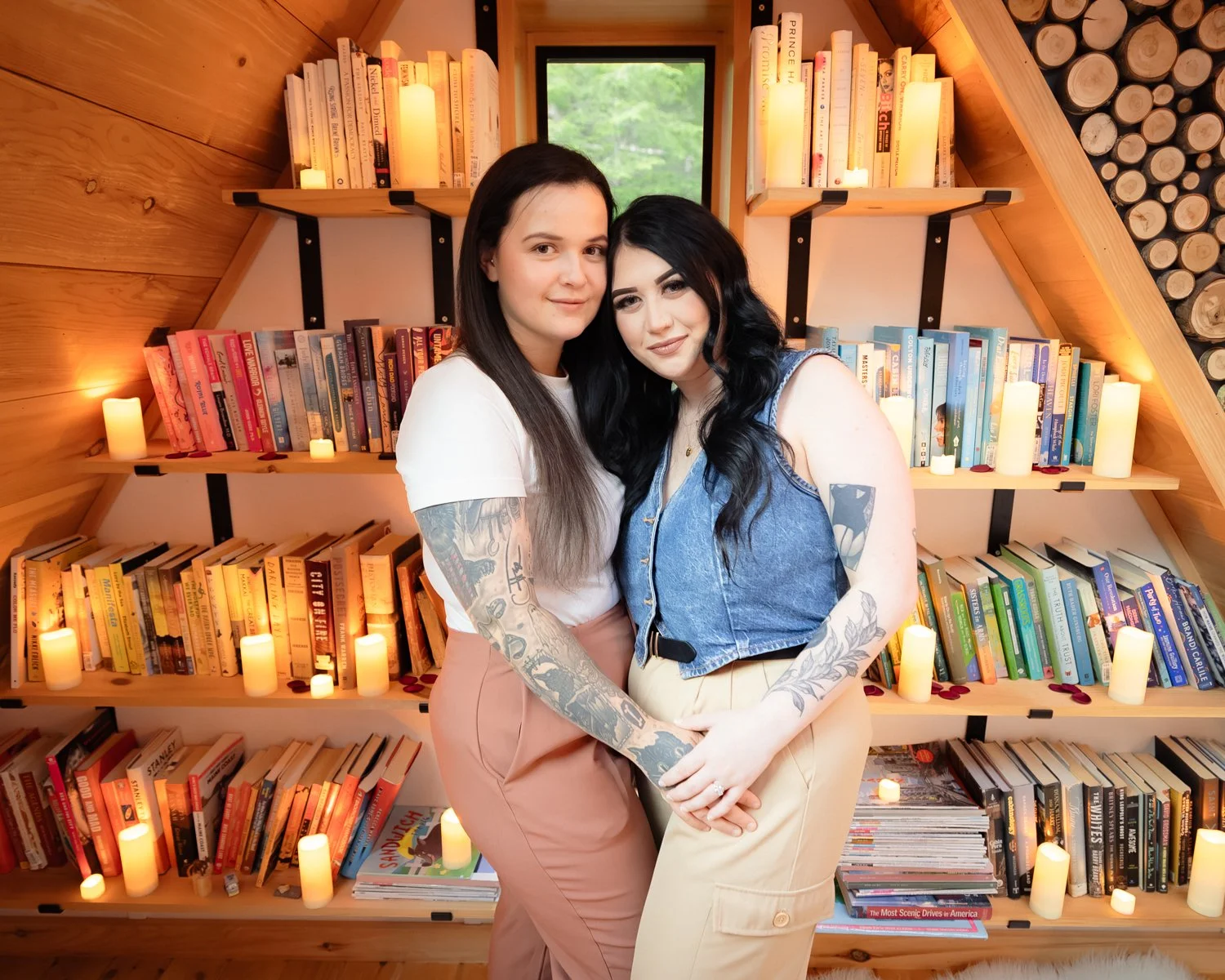 Two women standing close together in front of a wooden bookshelf filled with colorful books and decorative candles, inside a cozy, wood-paneled room with a small window showing greenery outside.