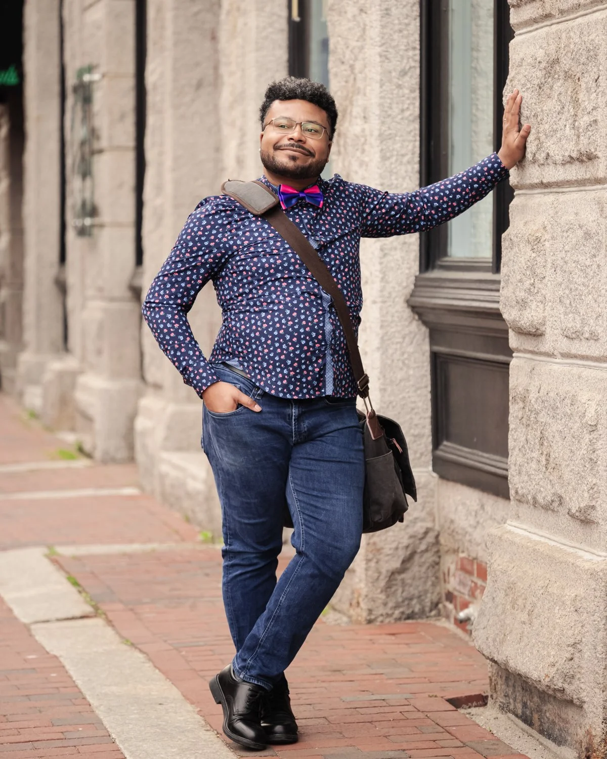 A man standing on a brick sidewalk, leaning against a stone building wall with one hand, wearing glasses, a patterned shirt, jeans, black shoes, a colorful bow tie, and carrying a shoulder bag.