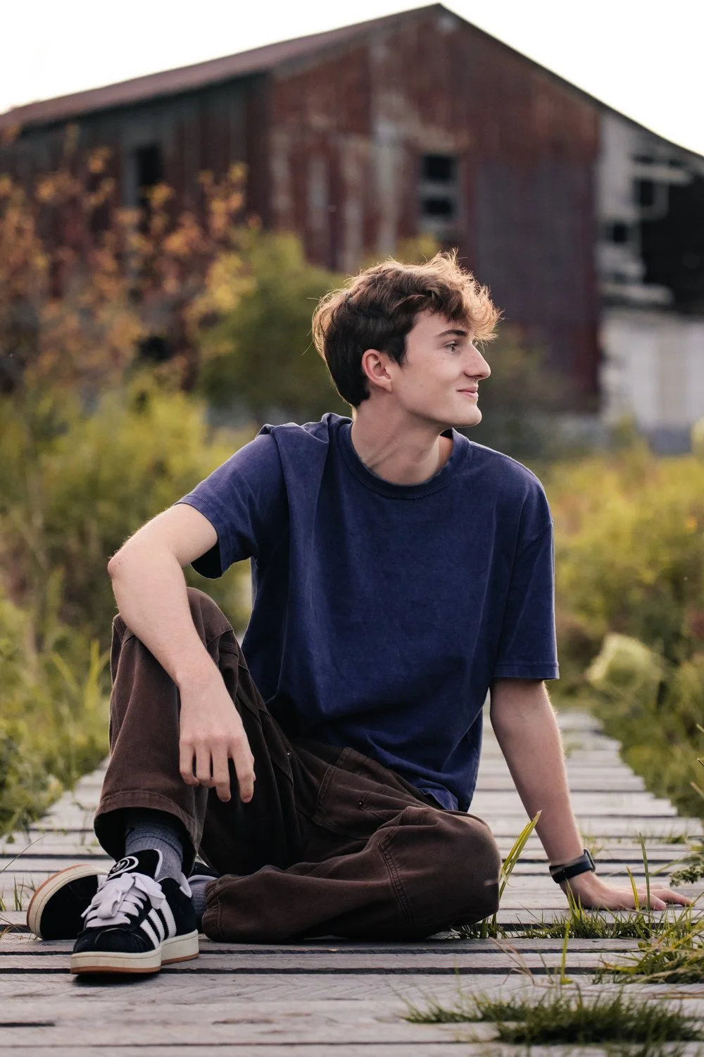 A young man with dark hair and a slight smile sitting on a wooden walkway outdoors, with a blurred barn and autumn trees in the background.
