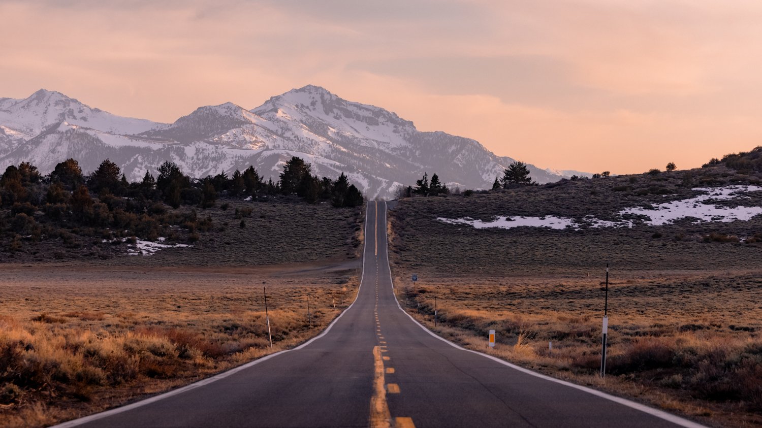 A long straight road stretching into the distance toward snow-capped mountains at sunset.