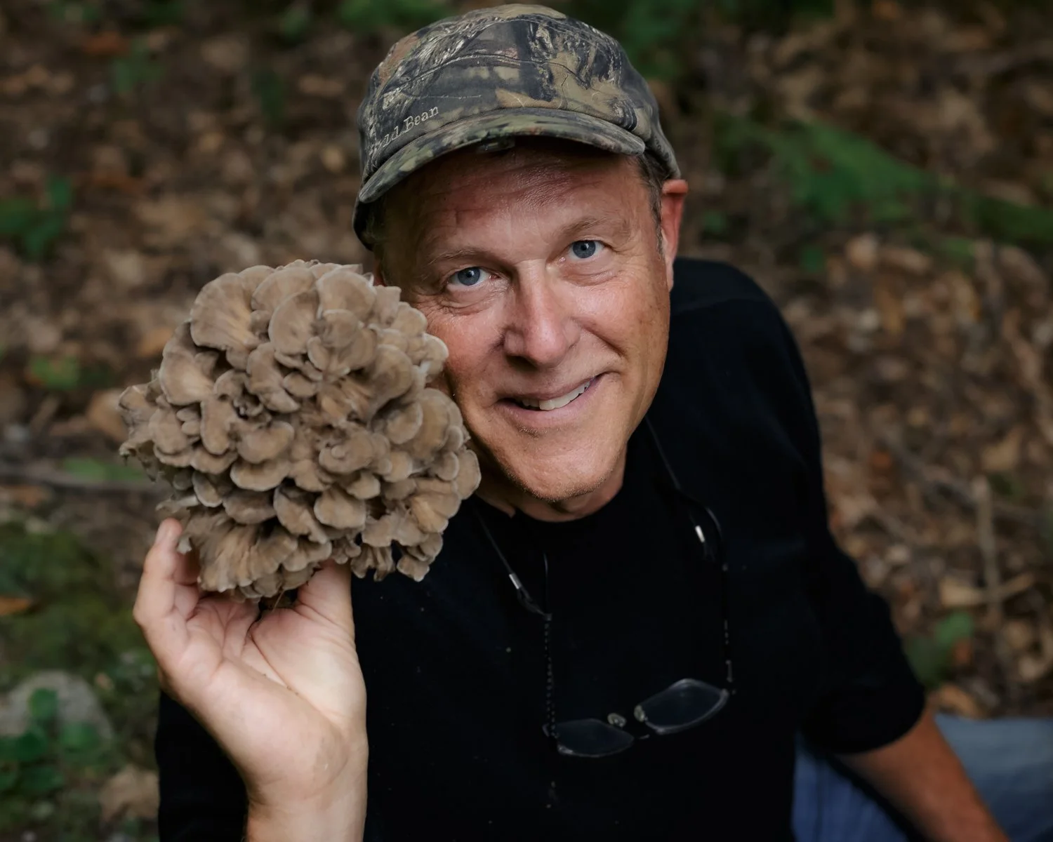 A man in a camouflage cap smiling and holding a large cluster of brown mushrooms in a forest.
