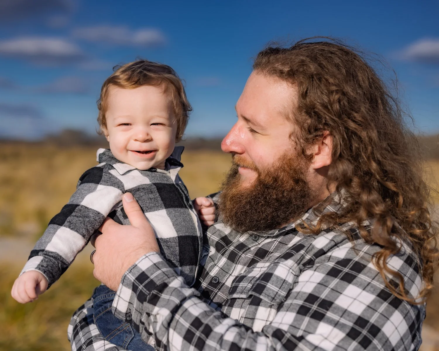 A man with long curly hair and a beard holding a smiling toddler outdoors on a sunny day.