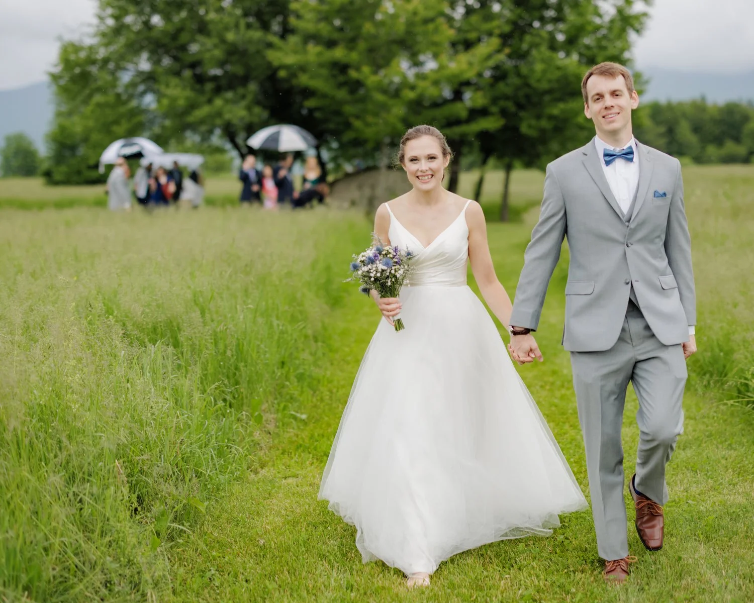 A newlywed couple walking hand in hand on a grassy field, with the bride in a white wedding dress holding a bouquet, and the groom in a light gray suit with a blue bow tie. In the background, people with umbrellas gather near a large tree.