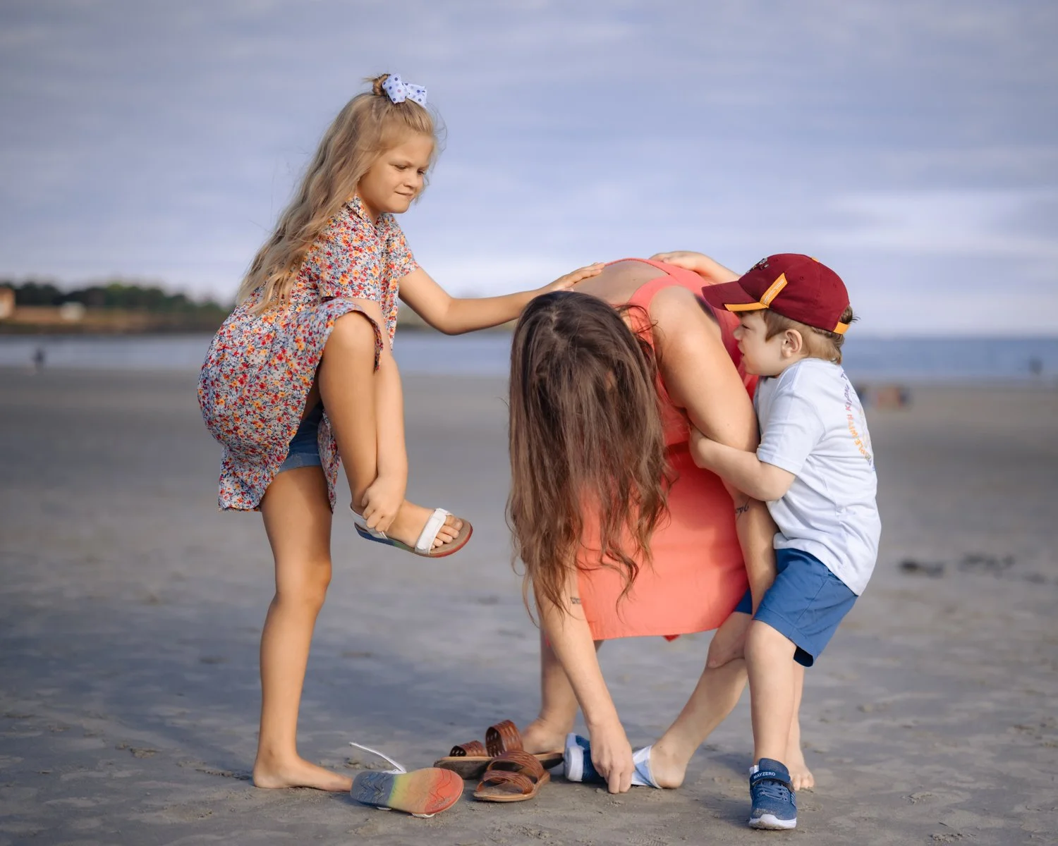 A woman with long brown hair standing on a beach, appearing distressed, with young children comforting her. One girl, with long blonde hair and a bow, is kneeling on the woman's back; a boy, wearing a white shirt and red cap, hugs her side.