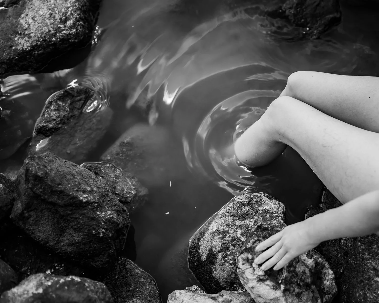 Child's legs and hand resting on rocks by a small stream or pond.