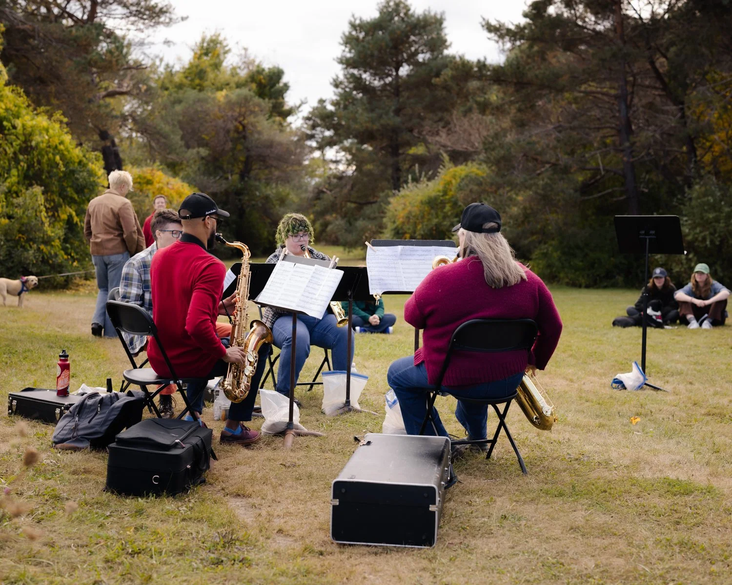 Group of musicians playing saxophones outdoors with sheet music in front of them, seated on chairs on a grassy field surrounded by trees, while some spectators sit on the grass nearby.