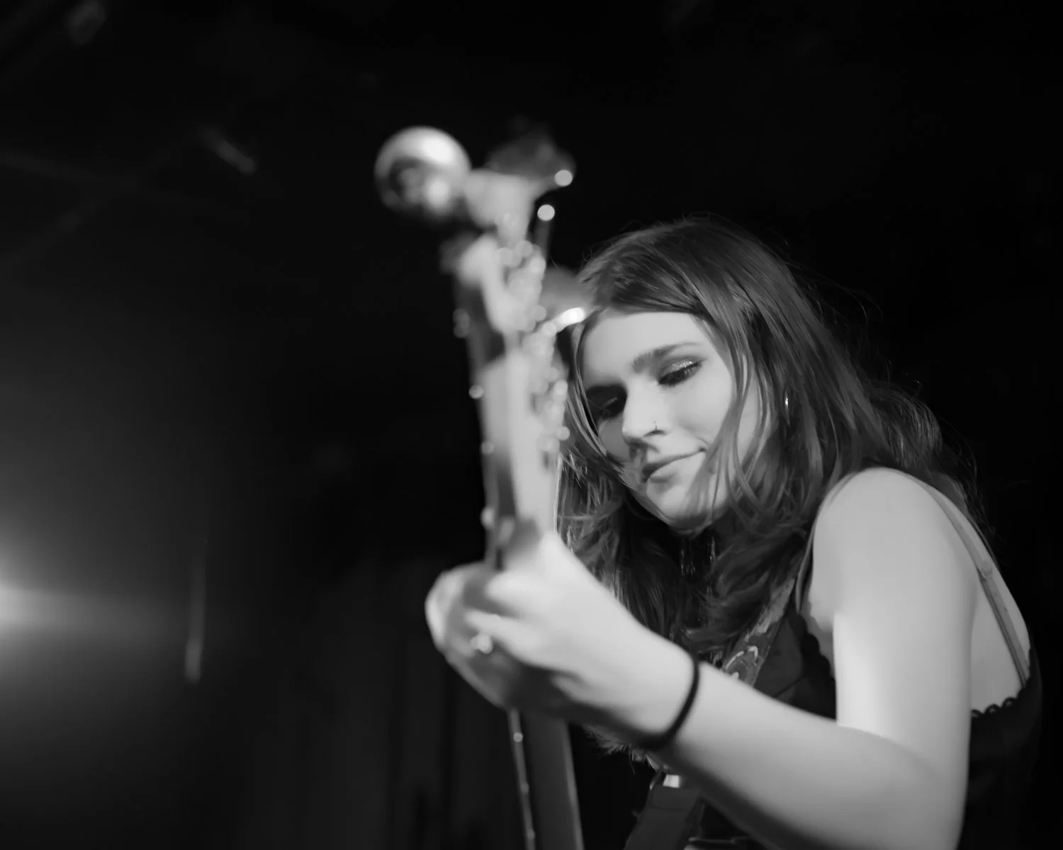 A young woman with wavy hair playing an electric guitar in a dark room, smiling softly.