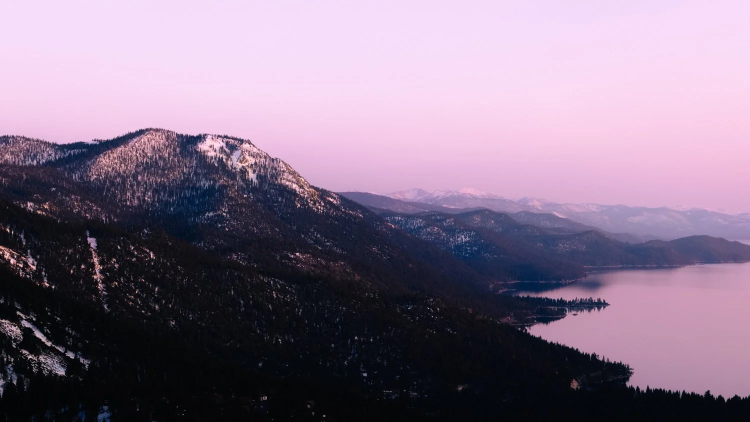 A mountain landscape at dusk with snow-capped peaks, forested slopes, and a calm lake reflecting the sky, which has a pinkish hue.