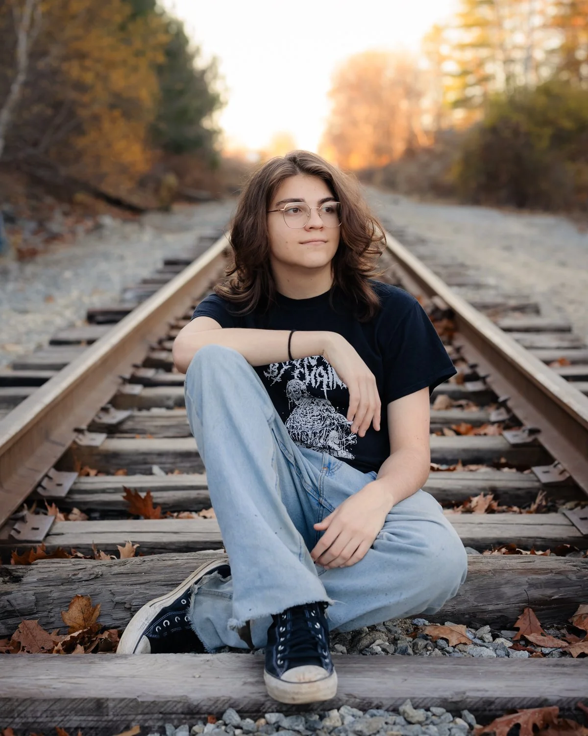 A young woman with glasses and long brown hair sitting on railway tracks surrounded by fallen autumn leaves, with trees and a sunset in the background.