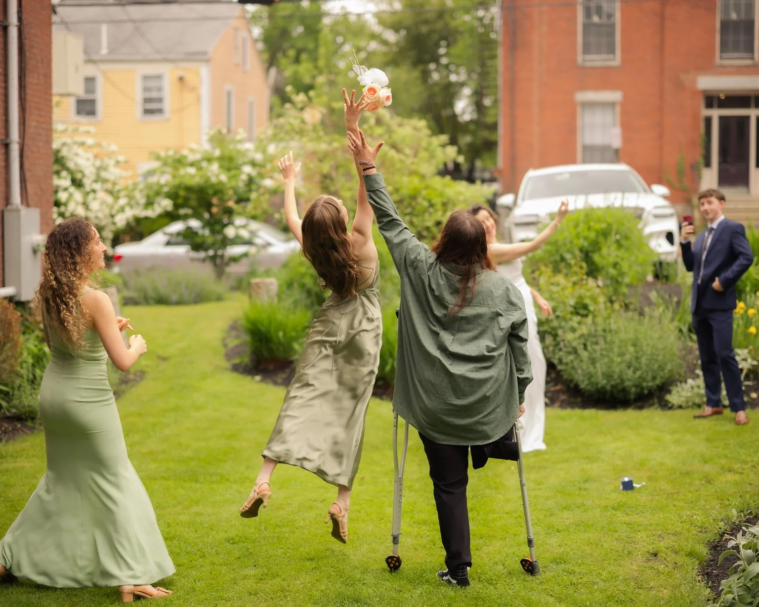 People celebrating outdoors in a garden with a woman in a white dress jumping, a woman on crutches reaching for a bouquet, others around them, in a residential neighborhood.