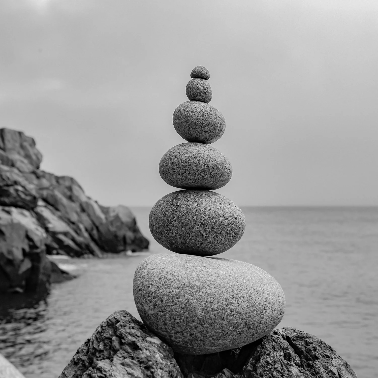 Stack of seven balanced stones on rocks near the ocean, black and white photo.
