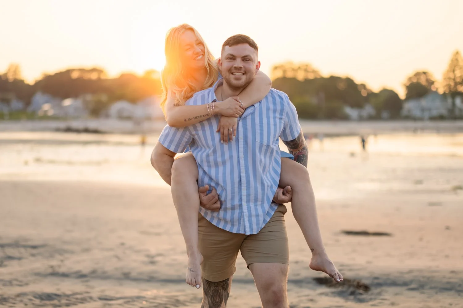 A couple at the beach during sunset. The woman is on the man's back, smiling and holding his shoulders. The man is smiling and supporting the woman. In the background, there are houses and trees with the sunset casting a warm glow.
