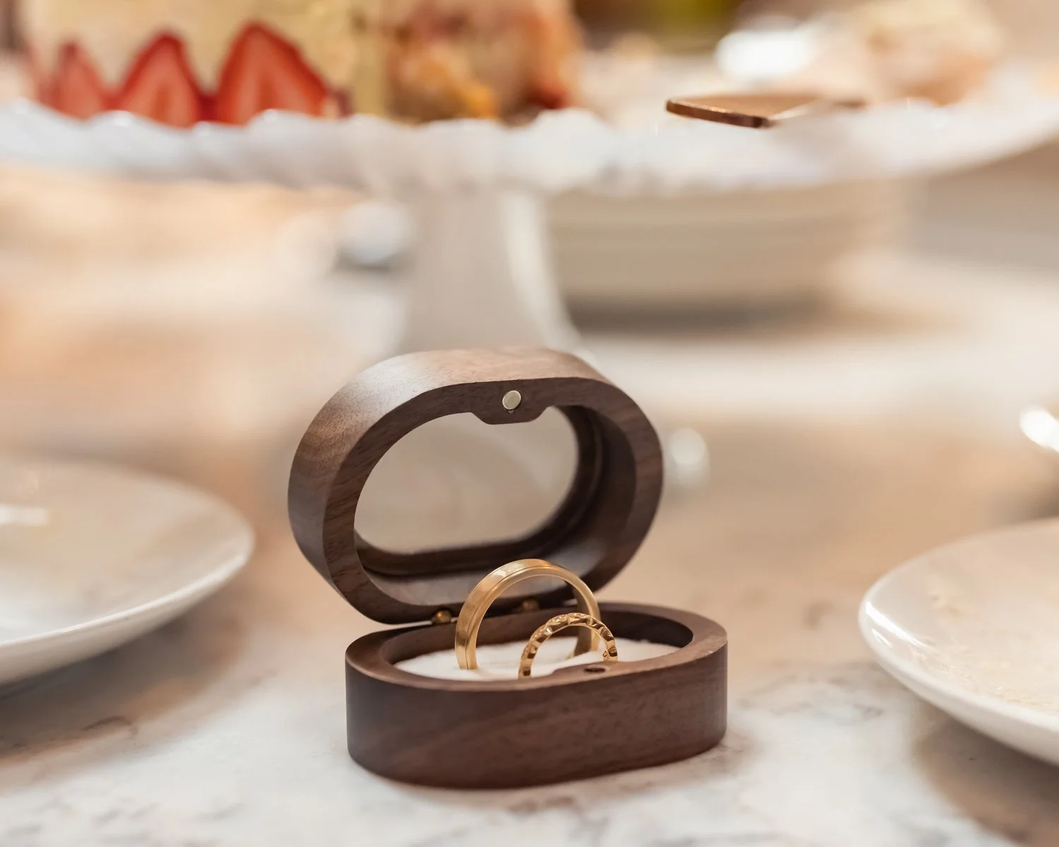 A wooden jewelry box with two gold rings inside on a table, with a blurry background of plates and strawberries.