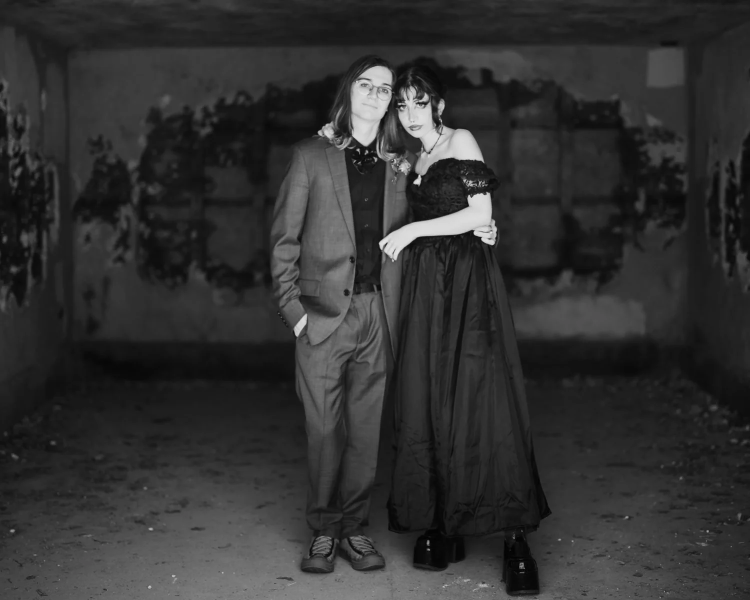 A man and woman stand together in a dimly lit, rustic space. The man is dressed in a plaid suit, black shirt, and sneakers, with one hand in his pocket. The woman wears an off-the-shoulder black lace top and a long black skirt with platform shoes. Th