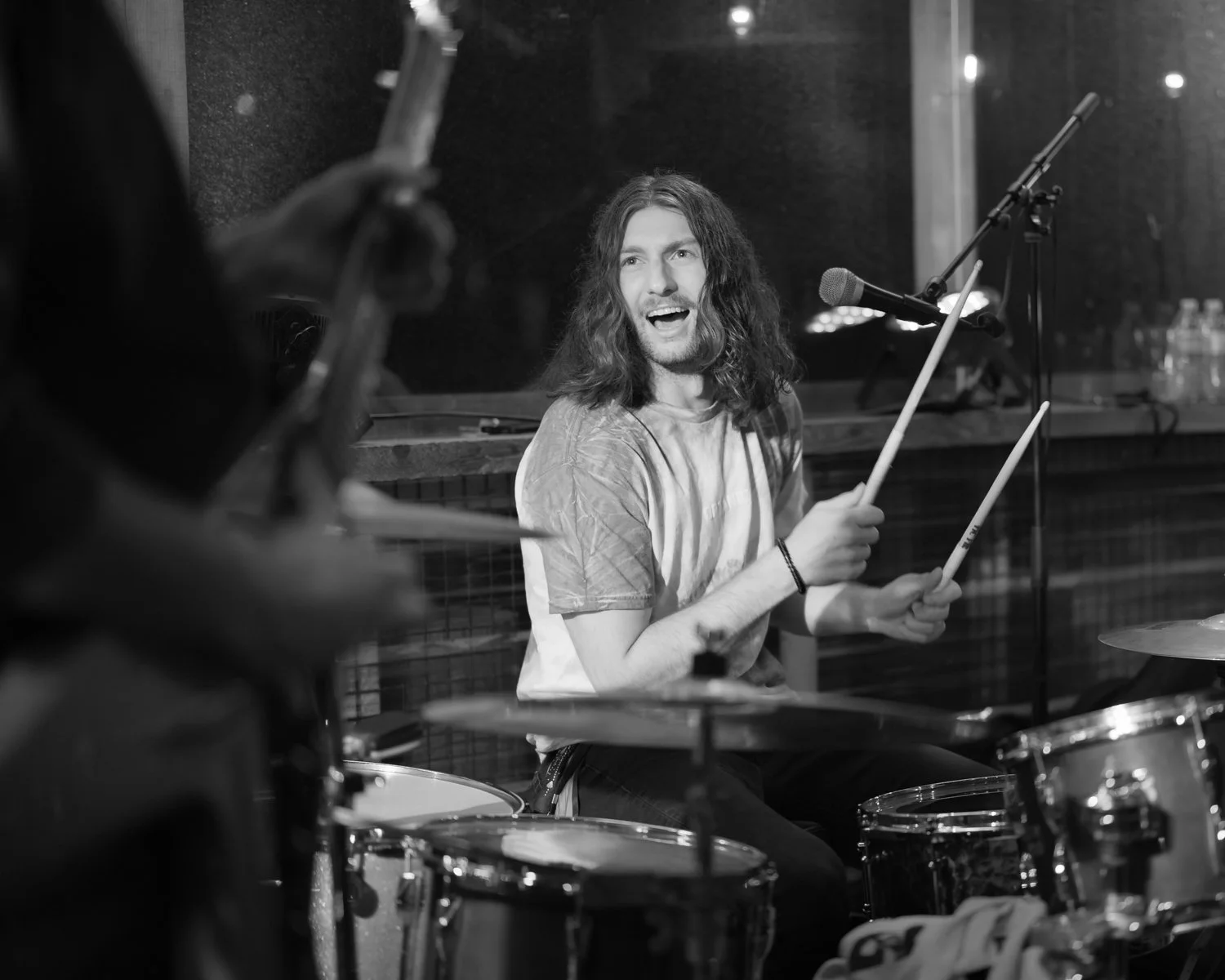 A young man with long curly hair playing drums during a performance in a music studio.