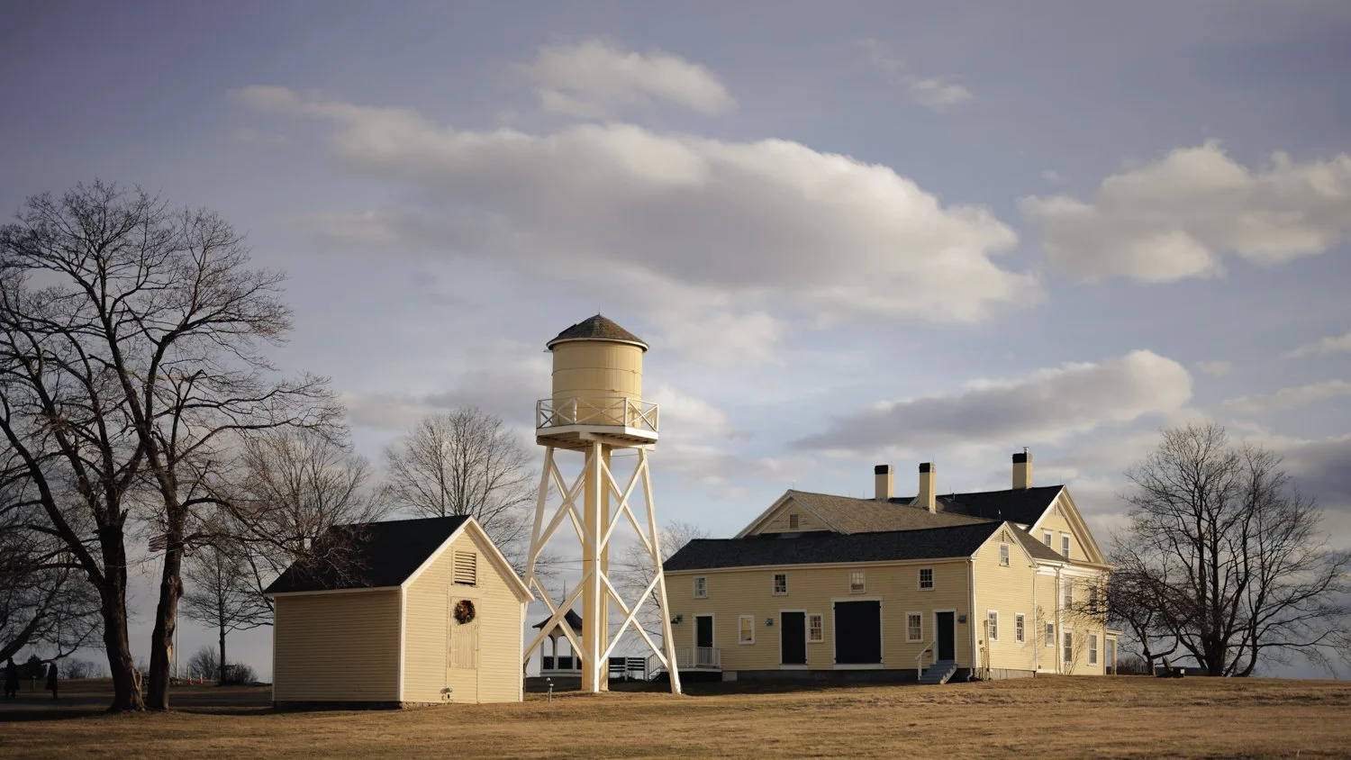 A yellow lighthouse with a small attached building and a large main building, all set in a grassy area with leafless trees and a cloudy sky in the background.
