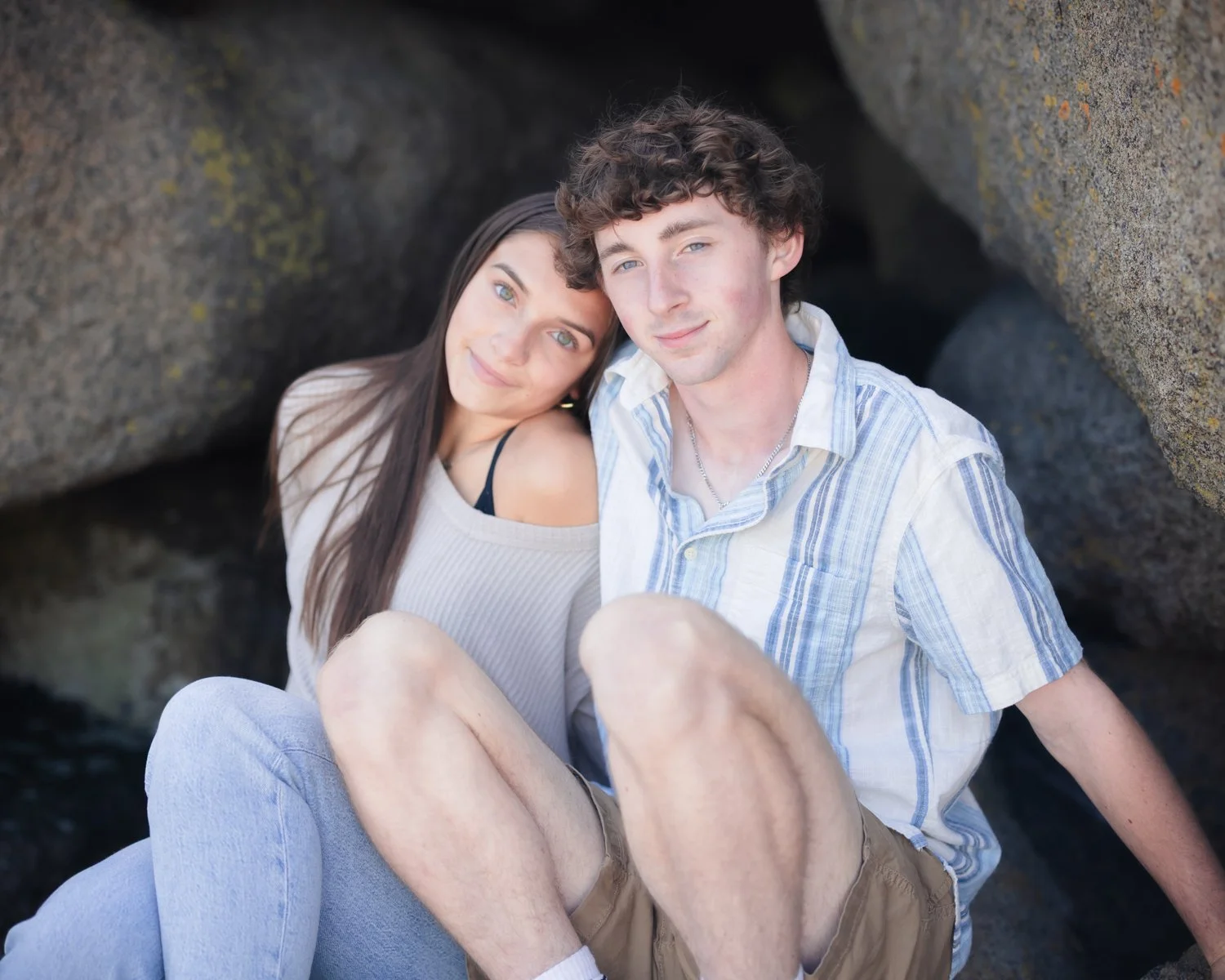 A young couple sitting together outdoors between large rocks, smiling at the camera.