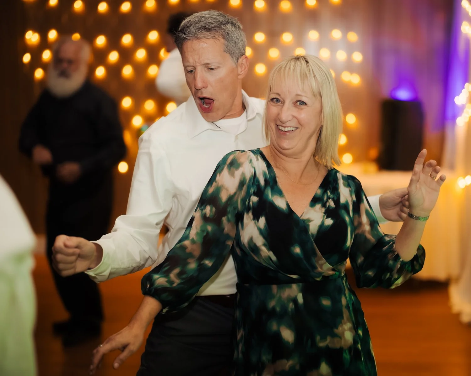 A man and woman dancing and smiling at a party or celebration with warm lights in the background.